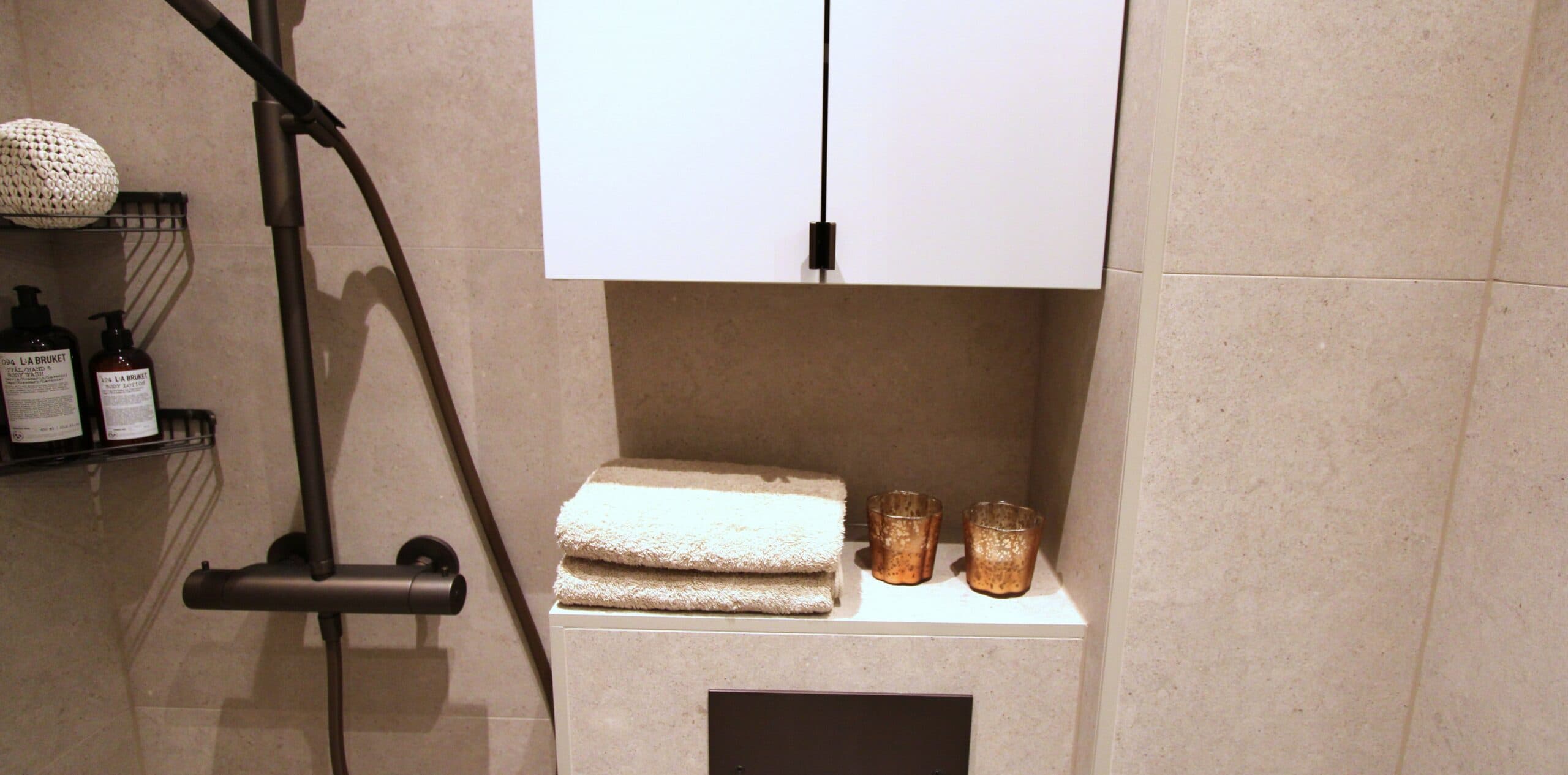 Närbild på ett modernt badrum med beige kakel, svart duscharmatur, vita skåpsluckor, vikta handdukar och två dekorativa ljuslyktor i kopparfärg.